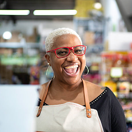 A woman with brown skin tones, red glasses, and white hair, wearing a black top, beige apron, and large teardrop earrings, smiles in a store.