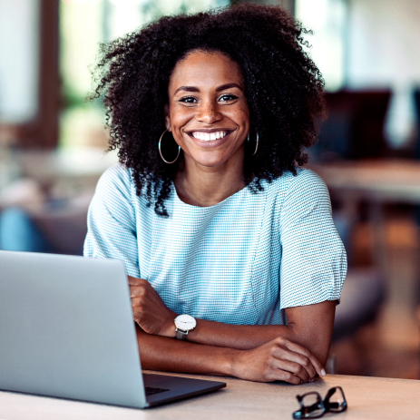 Uma mulher com tons de pele escuros e cabelos castanhos encaracolados sorri quando sentada em uma mesa com um laptop e seus óculos.